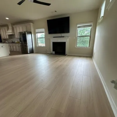 Spacious living room with light wood flooring, fireplace, mounted TV, black ceiling fan, and adjacent kitchen area.