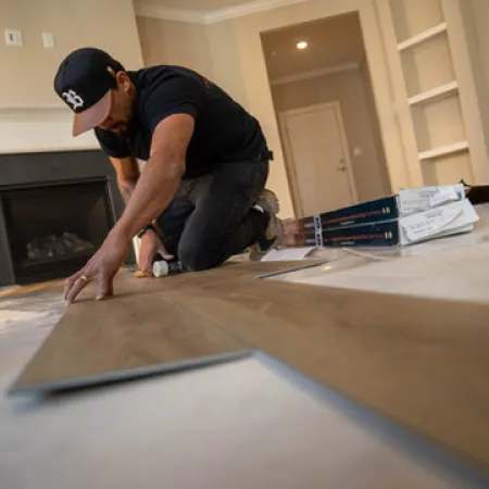 Man installing wood laminate flooring in a living room with fireplace and empty shelves.