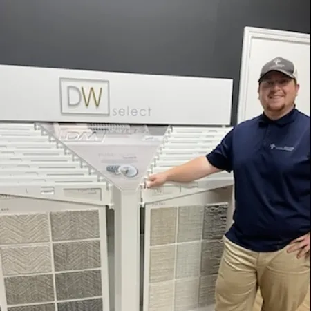 Man in navy shirt and khakis stands by DW Select carpet sample display with various textured carpet swatches.
