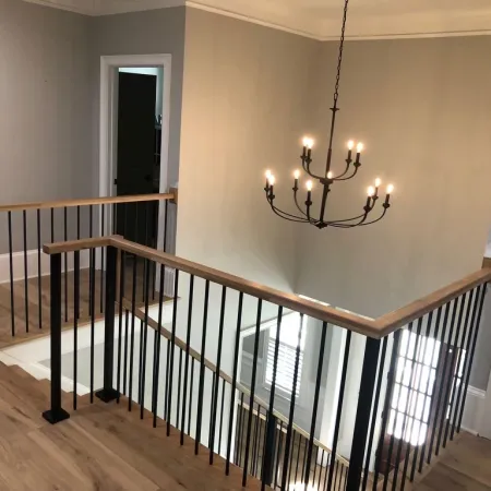 Modern staircase railing with black metal spindles and wooden handrail under a chandelier in a neutral hallway.