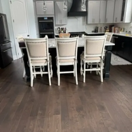 Modern kitchen with dark hardwood floors, white island with beige chairs, black appliances, and pendant lights.