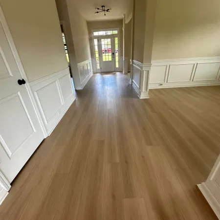 Empty hallway with light wooden floor, white wainscoting walls, and a glass front door letting in natural light
