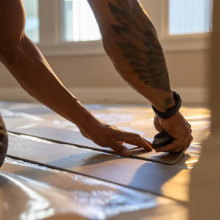 Man with tattooed arm using a tool to install wooden flooring in natural light.