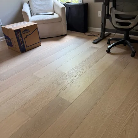 Bright home office corner with light wood flooring, white armchair, cardboard box, and black office chair.