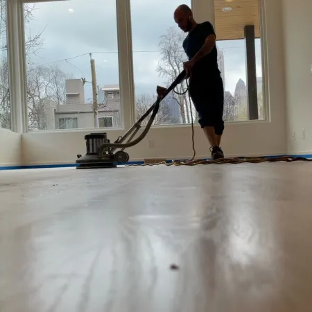 Man using floor polishing machine on wooden floor inside a bright room with large windows.