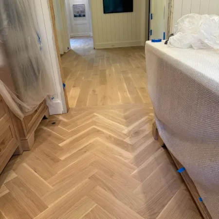 Light wood herringbone flooring transitions to plank flooring in a bright room with covered furniture and wall-mounted TV.