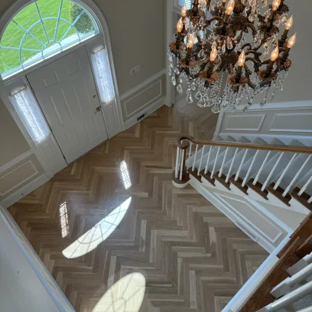 View of elegant foyer with wooden herringbone floor, crystal chandelier, and white front door with arched window.
