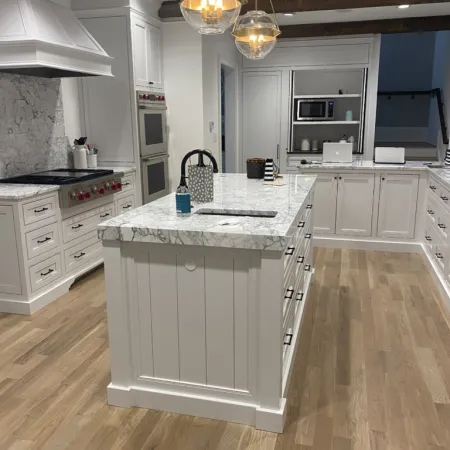Modern white kitchen with marble countertops, wooden floor, pendant lights, and stainless steel appliances.