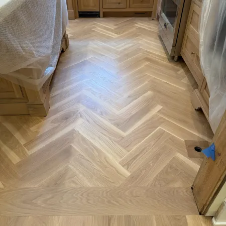 Light wood herringbone pattern flooring installed in a kitchen with natural wood cabinets and stainless steel stove.