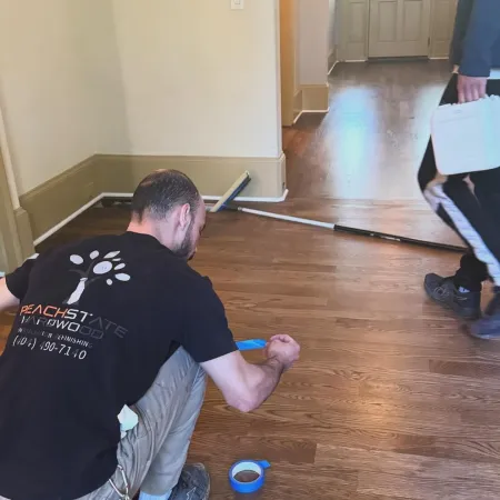 Worker taping hardwood floor during installation in a home with another person walking by holding a toolbox.