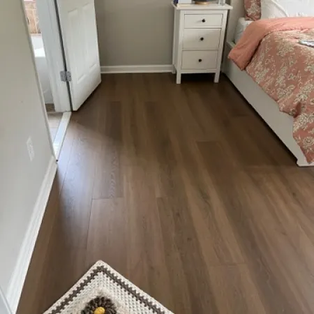 Cozy bedroom corner with floral bedspread, white nightstand, lamp, and wooden flooring with floral rug.