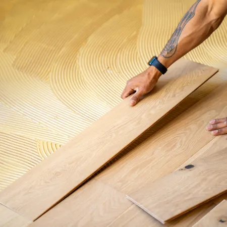 Hands installing wooden flooring over adhesive with textured patterns for a smooth fit and finish