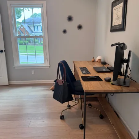Minimalist home office with wooden desk, computer monitor, office chair, window, and diploma on wall.