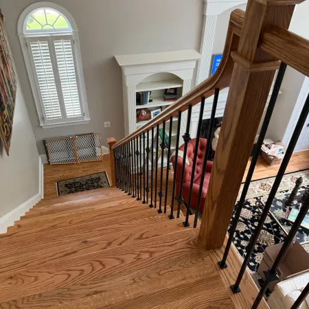 Interior view from top of wooden staircase with black metal balusters leading to living room with red chair and window.