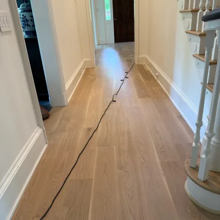 Long hallway with light wood flooring, white walls, staircase on the right, and a black cord running along the floor.