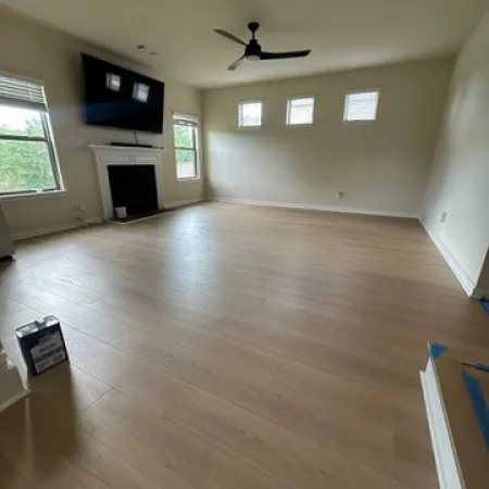 Empty living room with light wood flooring, ceiling fan, mounted TV, fireplace, and natural light from windows.
