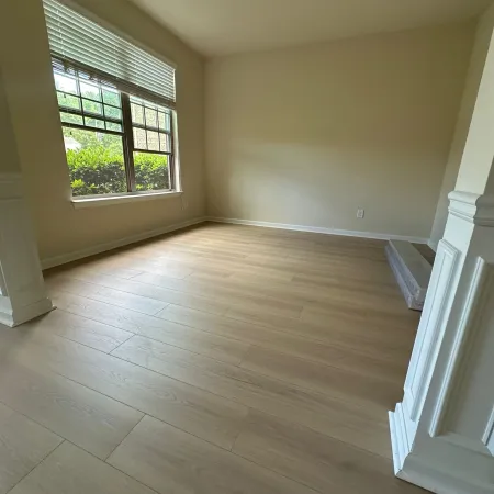 Empty room with light wood flooring, large window with blinds, beige walls, and modern ceiling light fixture.