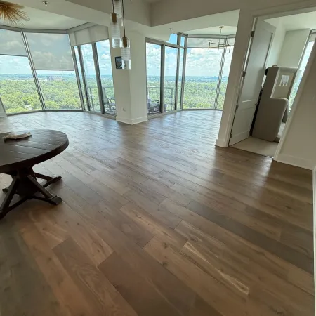 Spacious room with large floor-to-ceiling windows overlooking greenery and hardwood floors with round table.
