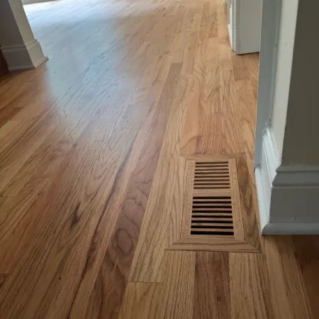Close-up of wooden floor with integrated floor vent near white baseboard and bright window in background