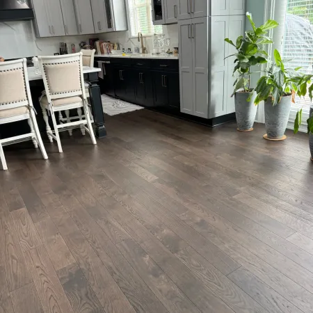 Modern kitchen with dark wood flooring, white and black cabinets, beige chairs, and potted plants by large windows