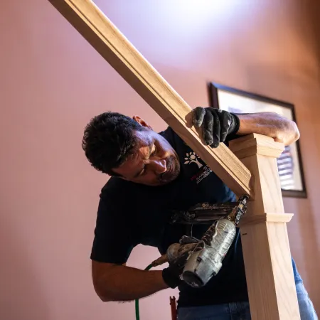 Man using nail gun to install wooden handrail on staircase inside a house with brown walls.