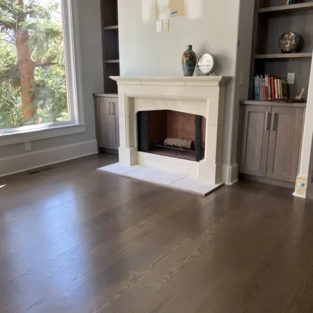 Bright living room with wooden floors, stone fireplace, built-in shelves, and large window showing trees outside.