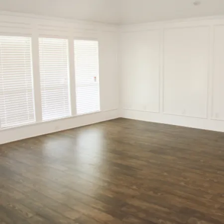 Bright empty room with dark wood flooring, white walls, large windows with blinds, and simple wall molding.