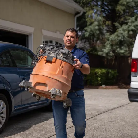 Man carrying large orange wet-dry vacuum cleaner on driveway between blue car and white van