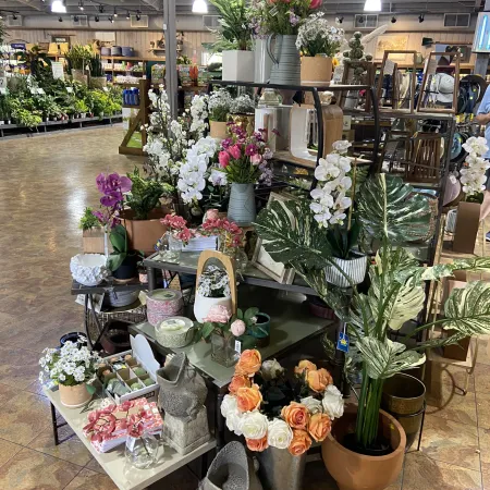Indoor display of artificial flowers, potted plants, and decorative items arranged on tables and shelves in a store.