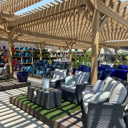 Outdoor wicker patio furniture with cushions under wooden pergola surrounded by colorful plant pots and garden decor