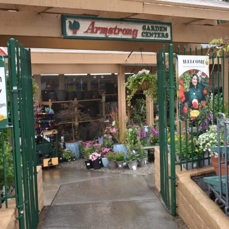 Entrance to Armstrong Garden Centers with green gates, plants, and signage showing open hours and welcome poster.