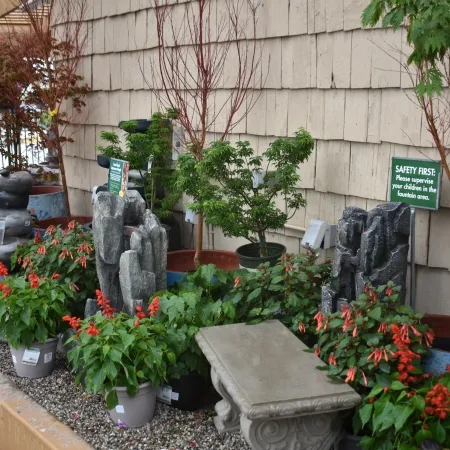 Outdoor garden display with green plants, red flowers, stone fountains, and a decorative stone bench near a beige wall.
