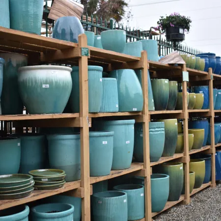 Rows of ceramic planters and pots in teal, blue, and green shades displayed on wooden shelves outdoors.