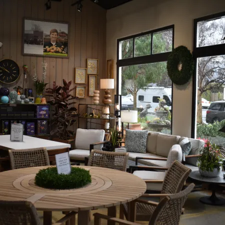 Cozy indoor seating area with wooden tables, wicker chairs, plants, and large windows overlooking cars outside.