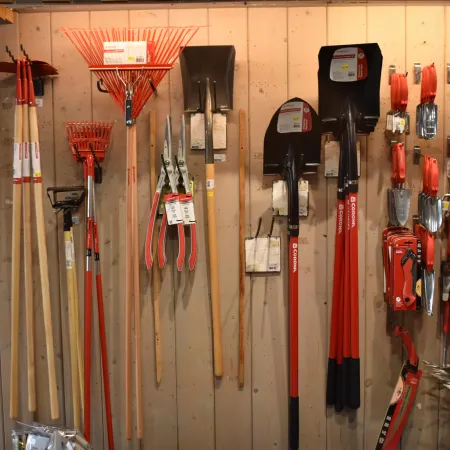 Garden tools including rakes, shovels, pruners, and hand tools hanging on a wooden wall in a store.