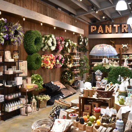 Cozy boutique store interior with floral wreaths, home décor, candles, and a lit PANTRY sign on wooden wall.