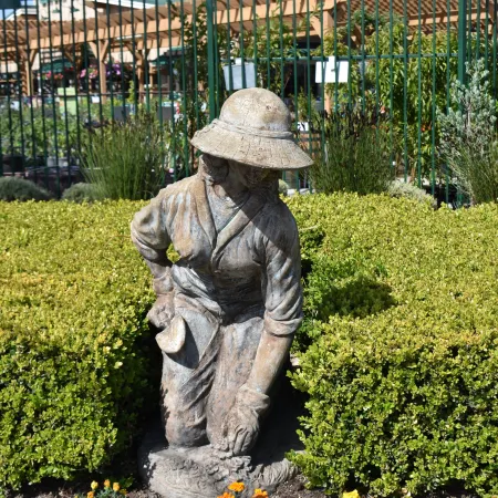 Stone statue of a gardener kneeling among yellow flowers and green bushes in a sunny garden area.