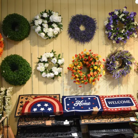 Colorful wreaths and patriotic-themed welcome mats displayed on a wall and shelf in a store.