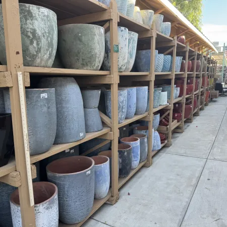 Rows of ceramic and stone plant pots displayed on wooden shelves in an outdoor garden center aisle.