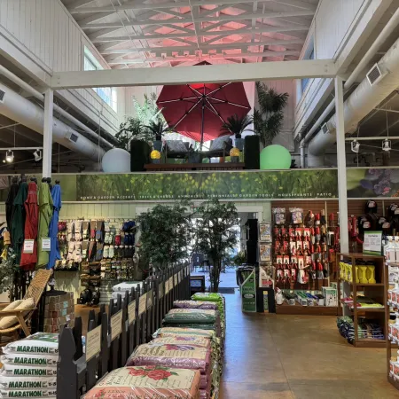 Interior of a garden supply store with outdoor furniture, gardening tools, bags of soil, and plants under skylights.