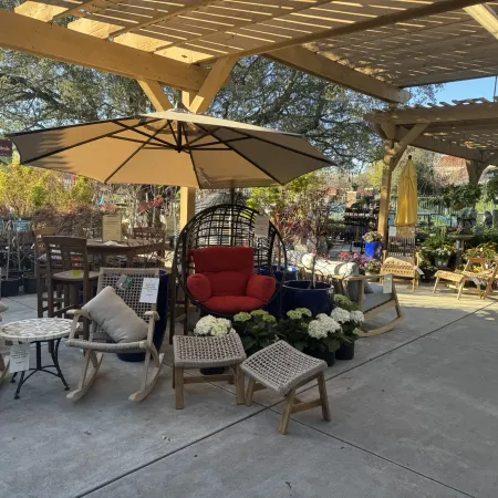 Outdoor patio furniture display with umbrellas, wicker chairs, a hanging chair, and potted plants under a wooden pergola.