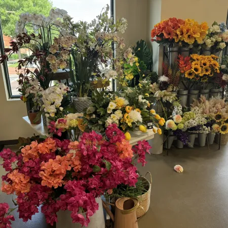 Colorful artificial flower bouquets arranged in a bright flower shop with large windows and natural light.