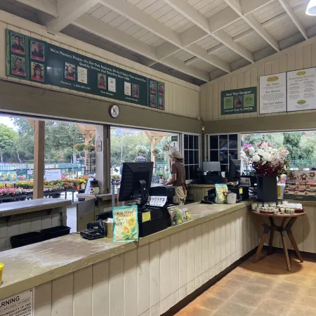 Bright garden center checkout with wooden counters, computer screens, flower displays, and a staff member assisting.