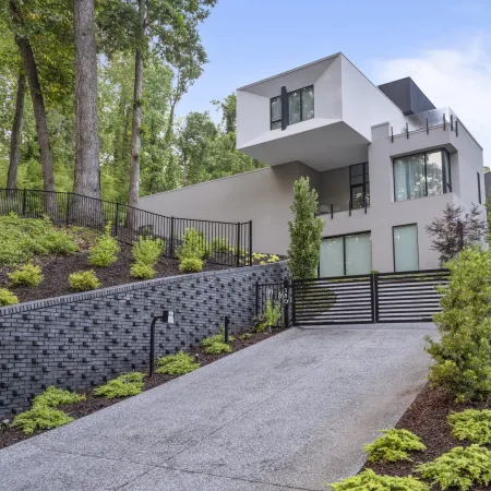 Modern multi-story home with cantilevered upper floor, landscaped yard, and sloped driveway with black fence gate.