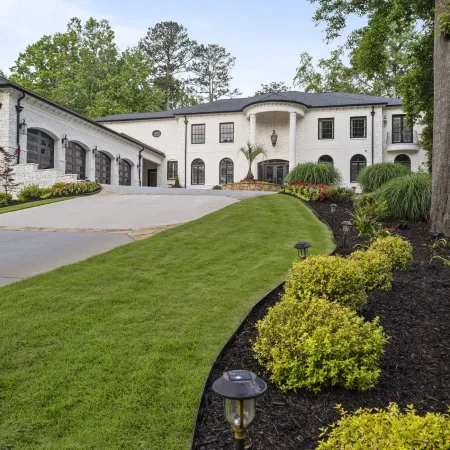 Large white two-story house with black roof, manicured lawn, garden beds, and driveway leading to a garage.