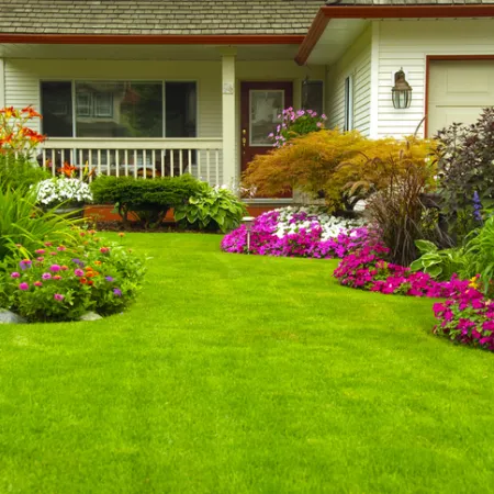 Front yard with lush green lawn and colorful flower beds surrounding a house porch with white railings.
