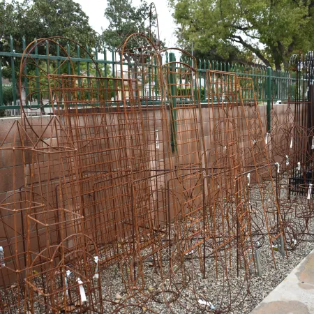 Rust-colored metal garden trellises of various shapes leaned against a wall on gravel near a sidewalk.