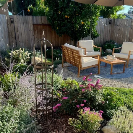 Sunny backyard garden with wooden fence, flowering plants, and outdoor seating area with cushioned chairs and table.