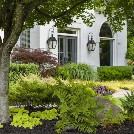 Lush green garden with various plants and a tree in front of a white house with black lanterns.