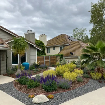 Front yard landscape with diverse plants, including purple and yellow flowers, palm trees, and stone pathways under a cloudy sky